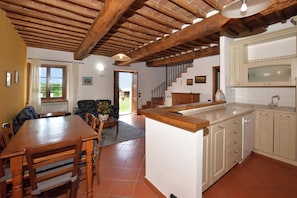 A spacious kitchen with wooden beams, a dining area with a table and chairs, and a view of the outdoors through a door. - Casa Sofi (Monticiano)