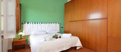 A bedroom with a green wall, wooden wardrobe, a bed with white linens, a nightstand with a lamp, and a window with a sheer curtain.