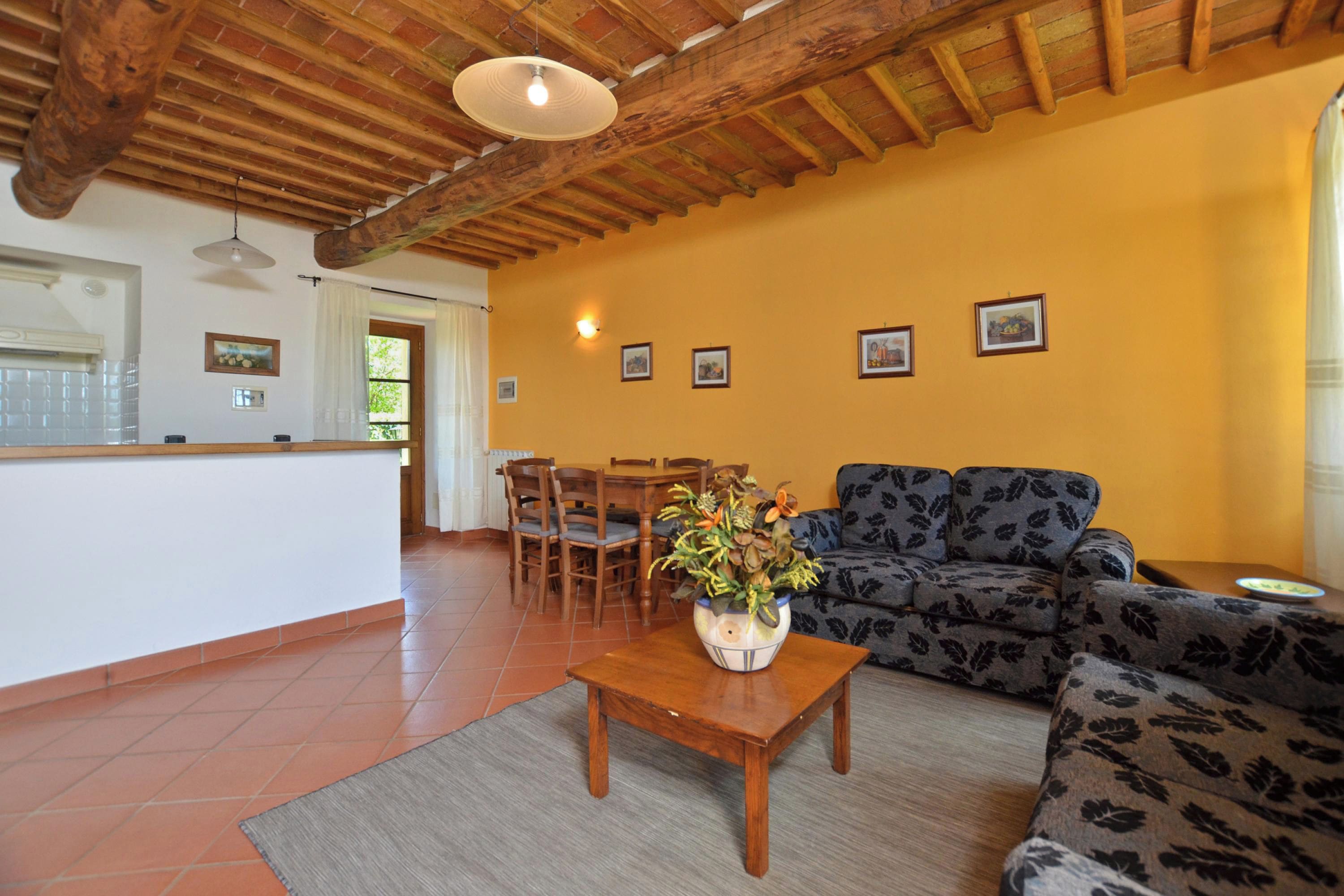 A living room with a sofa, a coffee table with a vase, a dining area with chairs, and a kitchen in the background.
