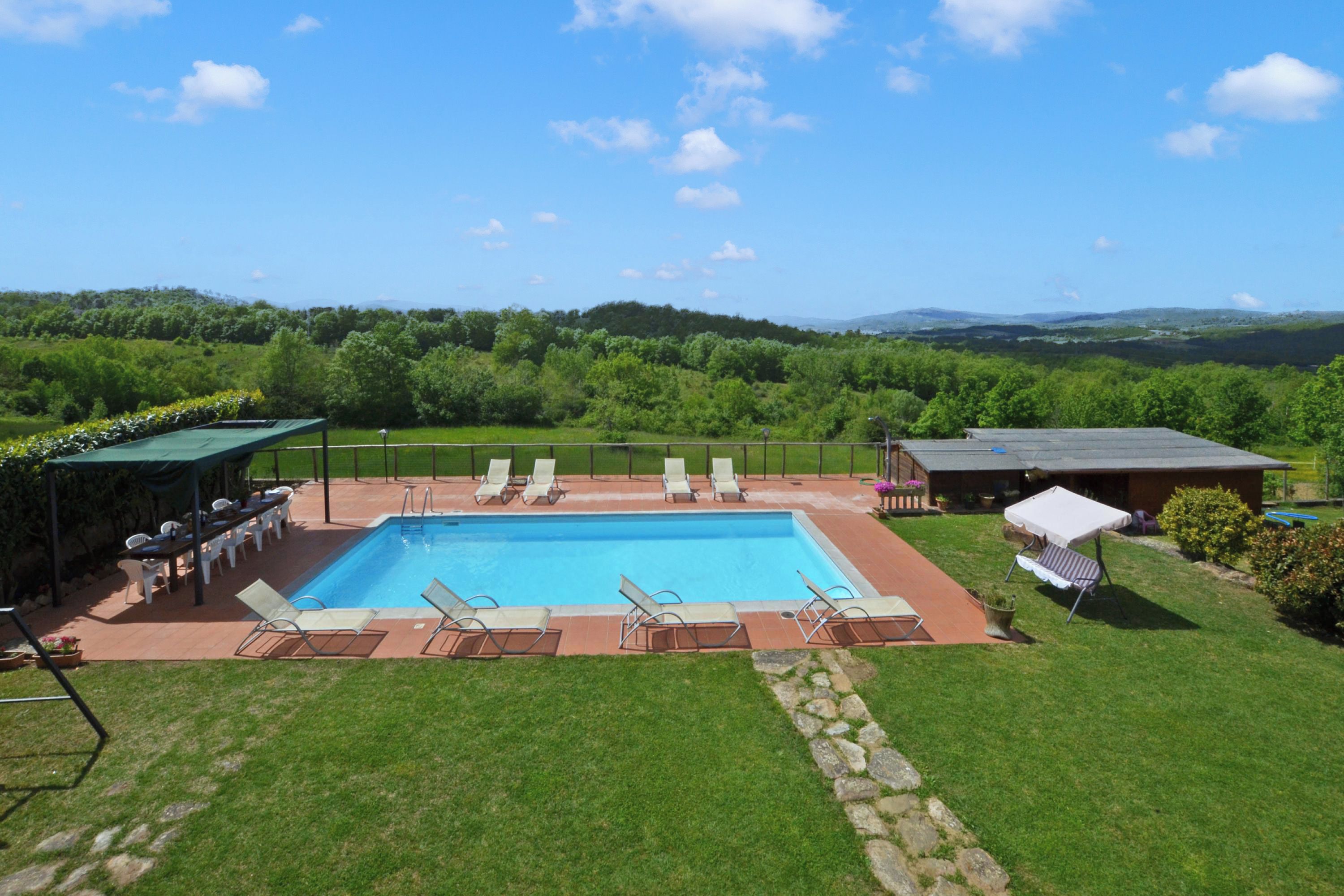 A rectangular swimming pool with lounge chairs and a covered seating area, surrounded by a well-maintained lawn and a view of a forested landscape.