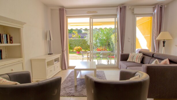 Ein Wohnzimmer mit Sofa, Sesseln, Couchtisch und Bücherregal. Ein Glastür verschiebt sich zu einem Garten mit Pflanzen und einem Tisch.