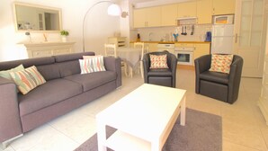 Ein modernes Wohnzimmer mit einem grauen Sofa, einem weißen Couchtisch und zwei Sesseln. Im Hintergrund ist eine Küche mit gelben Schränken und weißen Geräten zu sehen.