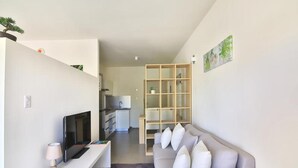 Un salón moderno con un sofá gris, un escritorio blanco y un televisor. Se observa una cocina al fondo con un armario de madera y una planta sobre la encimera.