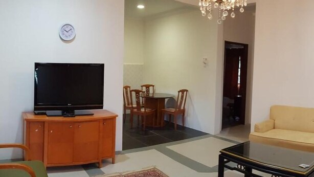 A living room with a television on a wooden cabinet, a beige sofa, a coffee table, and a dining area with chairs and a table. - Mri Homestay Sg Buloh - 3 Br House on First Floor With Centralised Pool (Sungai Buloh)