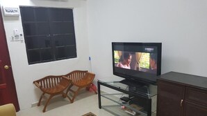 A living room with a television, two wooden chairs, a glass coffee table, a patterned rug, a cabinet, and a window with blinds.