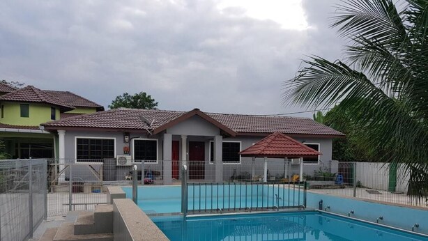 A swimming pool with a tiled deck, surrounded by a fence, and adjacent to a building with a red roof and white walls.