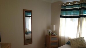 Un dormitorio con cama, cómoda, espejo, mesita de noche con lámpara y cortinas.