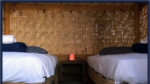 A room with two wooden beds, a small wooden nightstand with a pink lamp, and a woven bamboo wall.