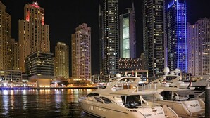 Een jachthaven met aangemeerde jachten, een stadsachtergrond met verlichte gebouwen, en een hotel genaamd "RAMADA" op de achtergrond.