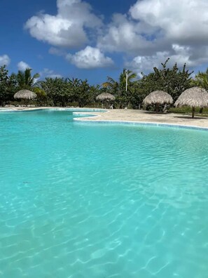 A clear turquoise pool with a sandy beach and thatched umbrellas. - Bright and Beautiful Aapartment -bavaro, Punta Cana (Punta Cana)