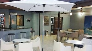 Un espace bar avec des chaises et des tables blanches, un parasol blanc et une vue sur la mer.