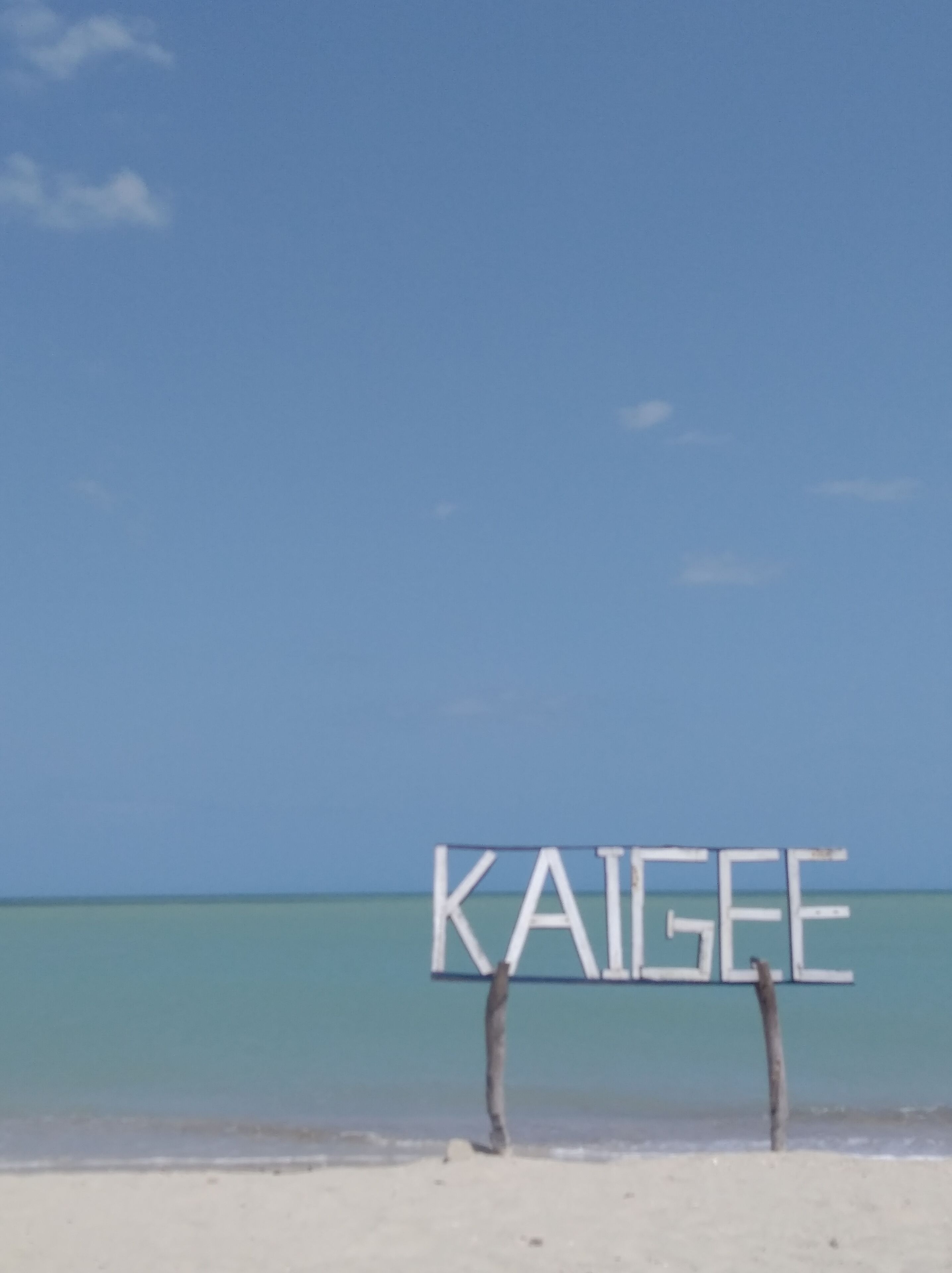 Segnaletica da spiaggia con la scritta "KAIGE" su una spiaggia sabbiosa e acque blu cristalline.