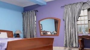 A bedroom with a wooden bed, a nightstand, a lamp, a mirror, and a window with curtains. - Room-christine Apartments-guest Room (Port Harcourt)