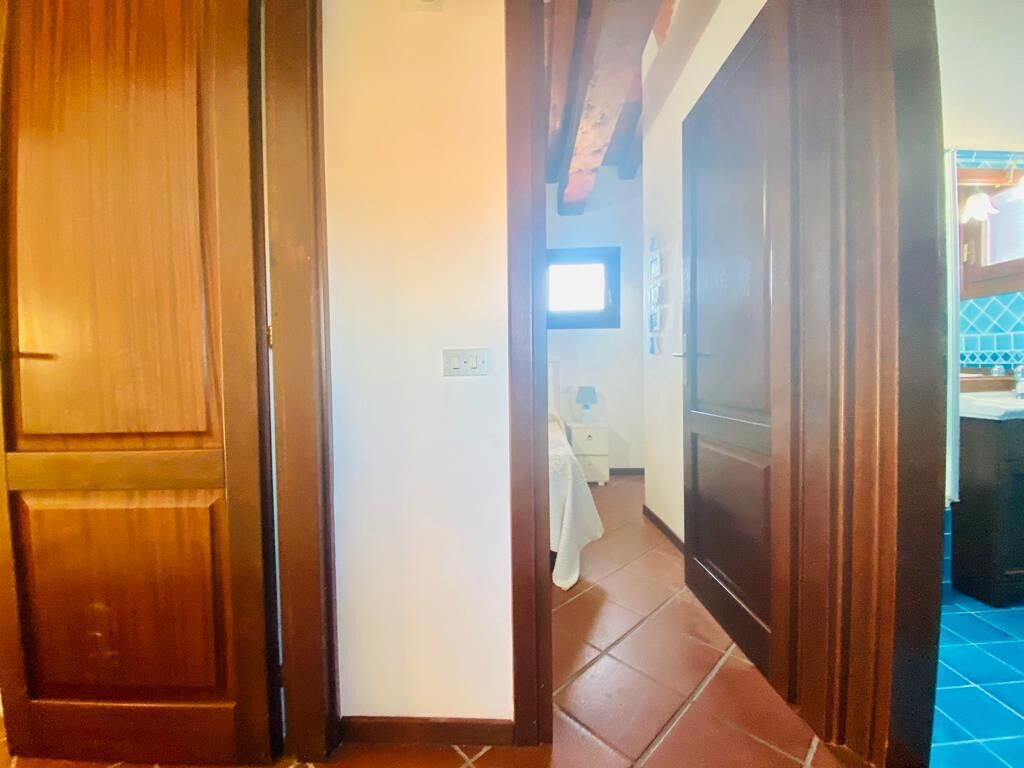 A hallway with wooden doors and tiled flooring leading to a bedroom with a bed and a nightstand.