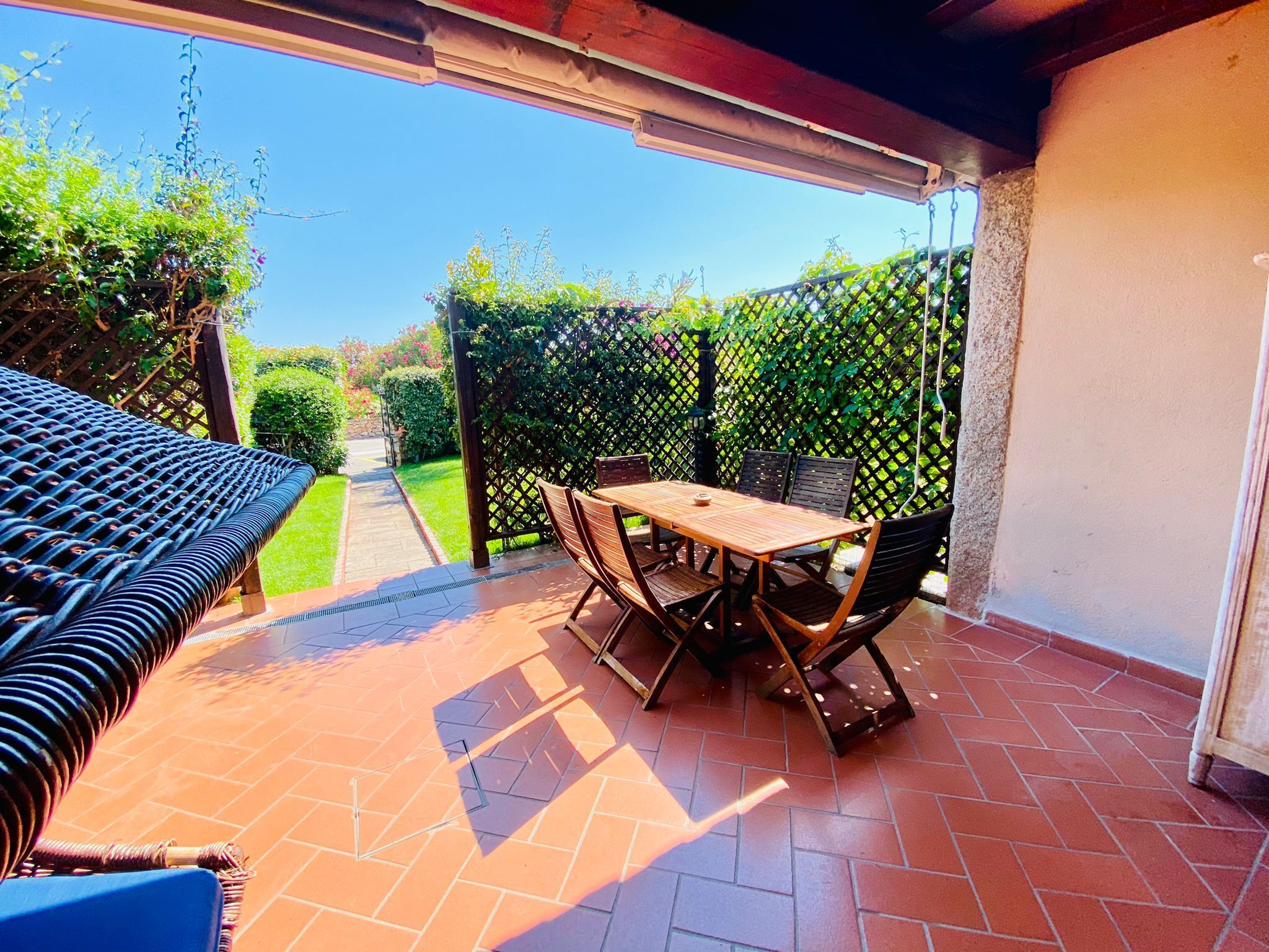 Une terrasse avec une table et des chaises, un canapé en osier et une vue sur un jardin, visible à travers une clôture en treillis.