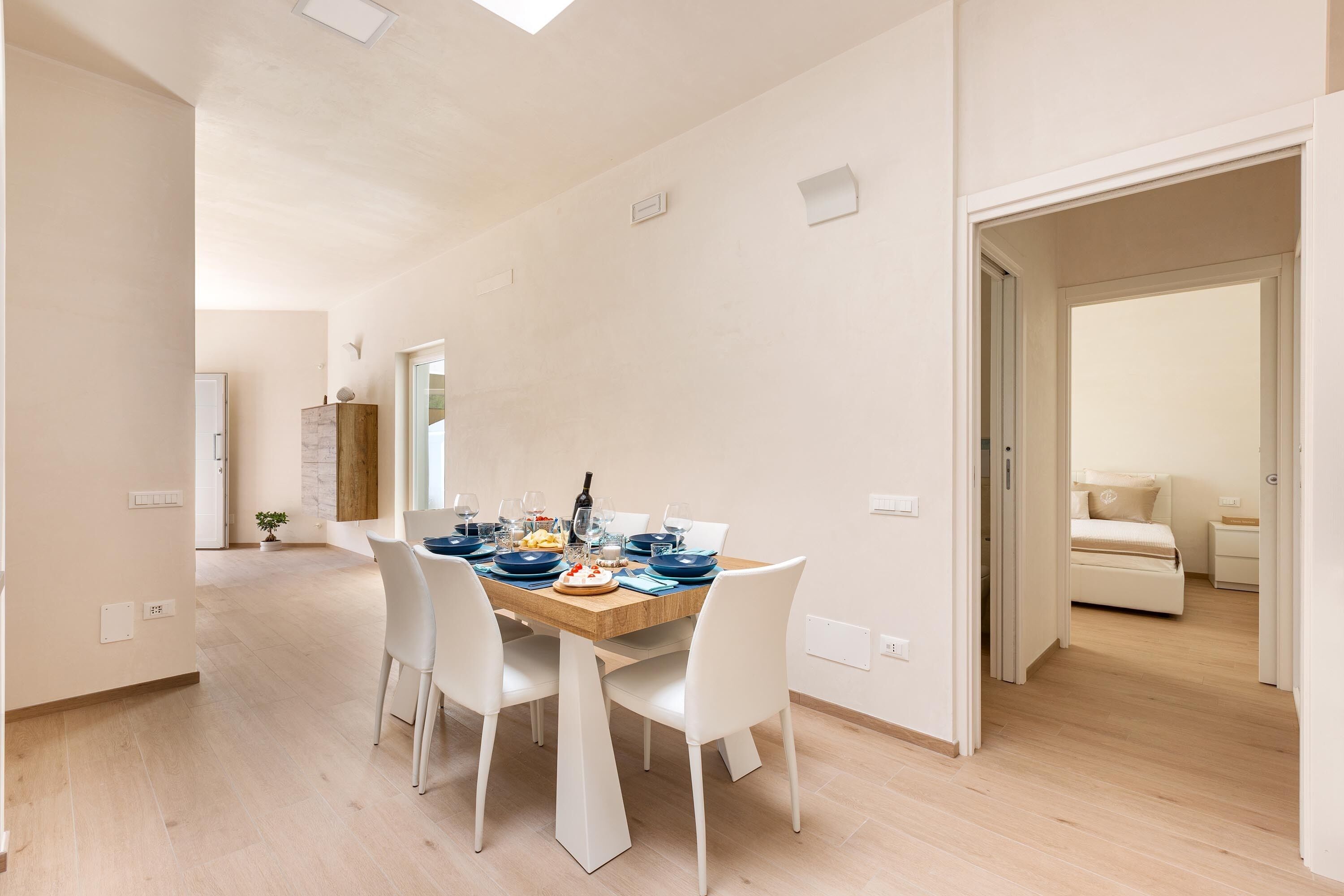 A spacious dining area with a wooden table set for four, white chairs, and a view into a bedroom with a bed and nightstand.