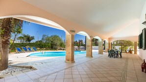 A covered pool area with tiled flooring, blue lounge chairs, and a table with chairs. - 1929 Villa Simona (Ugento)