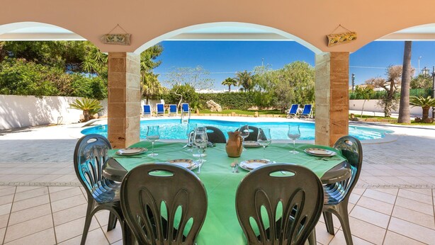 An outdoor dining area with a table set for four, overlooking a swimming pool and garden. - 1929 Villa Simona (Ugento)