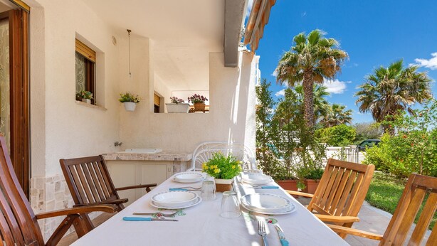 A dining table set for four with white tablecloths, wooden chairs, and a view of palm trees and greenery. - 1965 Villino dei Pini (Galatone)