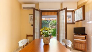 A dining area with a wooden table, chairs, a vase with flowers, a television on a stand, and a view of greenery through an open door.