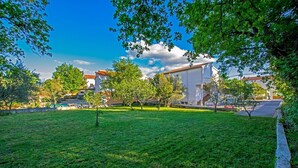 Un cortile erboso con alberi, un edificio bianco e un cielo azzurro limpido.
