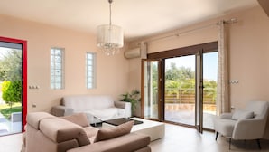 A modern living room with a sofa, armchair, and a coffee table. There is a sliding glass door leading to a balcony with a view of greenery.