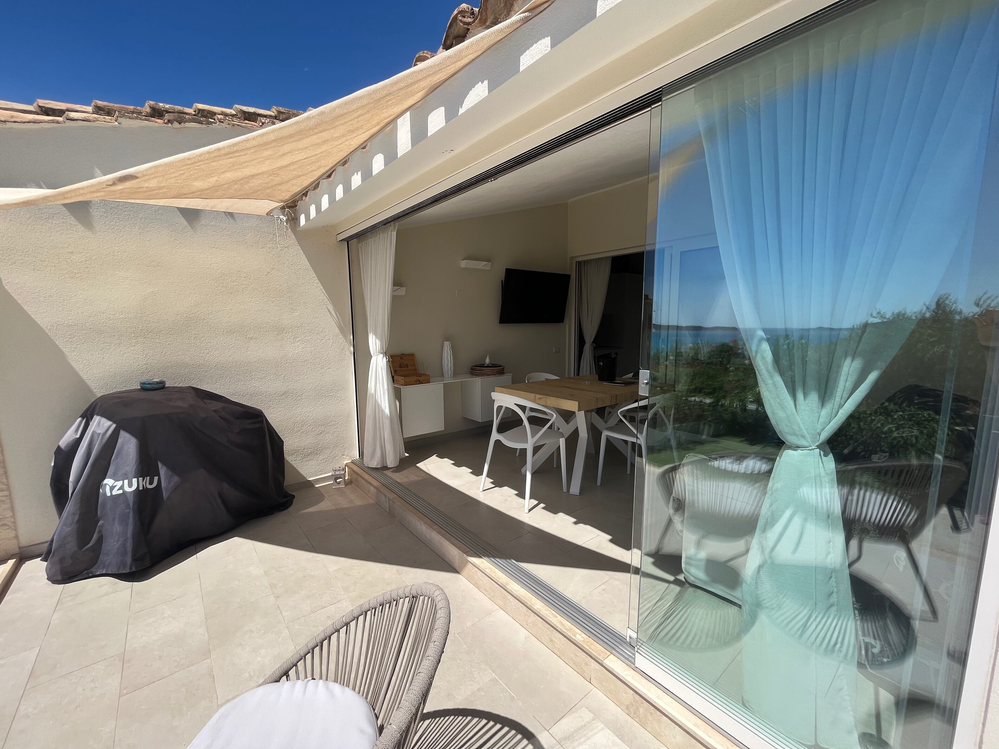 Ein Balkon mit Grill, Tisch, Stühlen und einer Glastür, die zu einem Innenbereich mit Fernseher führt.