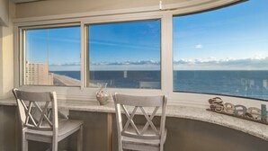 Quarto com duas cadeiras brancas e uma janela curva com vista para a praia e o oceano.