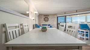 Uma sala de jantar com uma grande mesa de madeira, cadeiras brancas, um quadro de leme de barco na parede e vista através de grandes janelas.