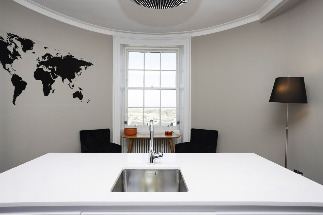 A modern kitchen with a white countertop, a stainless steel sink, a window with a view, a world map on the wall, and a floor lamp.