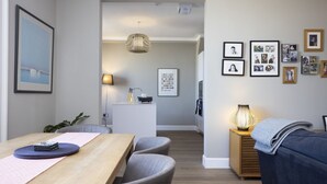 A modern living room with a wooden dining table, grey chairs, a blue sofa, and a small side table with a lamp.