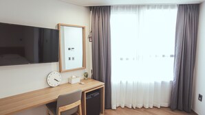A hotel room with a wooden desk, a chair, a television, a clock, a small plant, and a window with curtains.