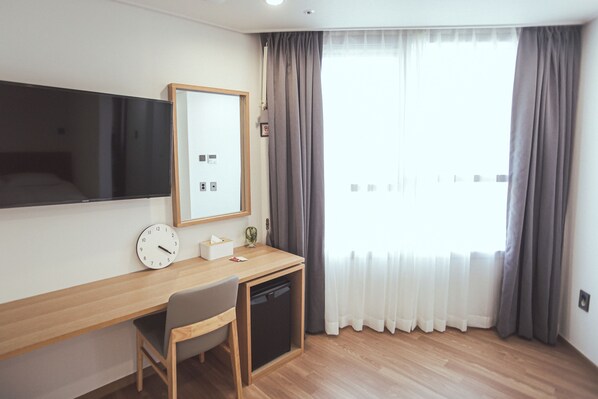 Ein Hotelzimmer mit einem Holztisch, einem Stuhl, einem Fernseher, einer Uhr, einer kleinen Pflanze und einem Fenster mit Vorhängen.