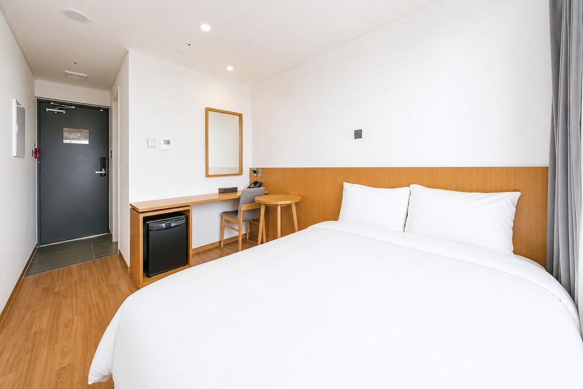 A hotel room with a bed, a desk, a chair, a small refrigerator, and a mirror.