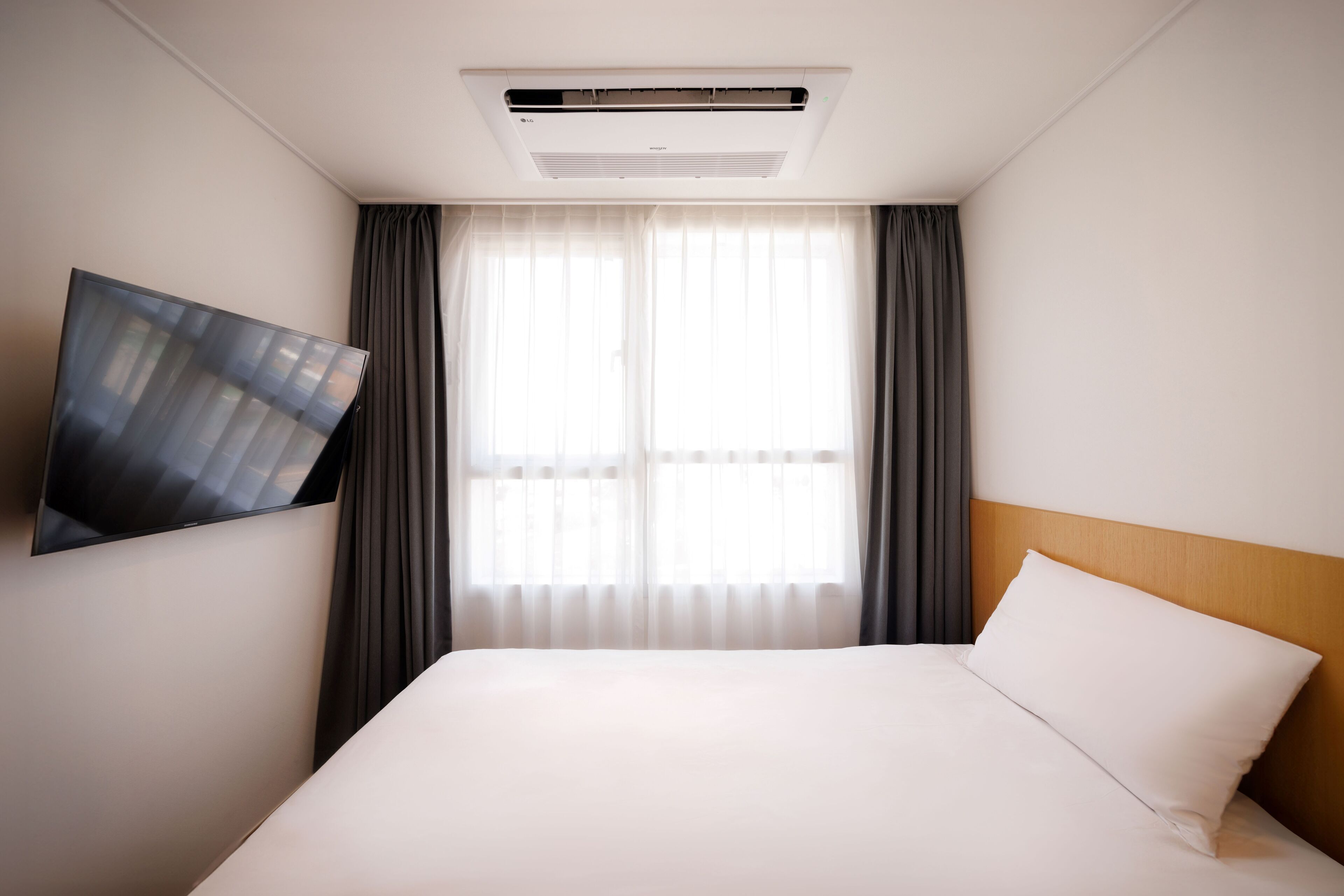 A hotel room with a bed, a mounted flat-screen TV, a window with curtains, and an air conditioning unit.