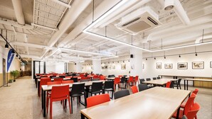 A spacious conference room with multiple tables and chairs, white walls with framed pictures, and exposed ceiling pipes.