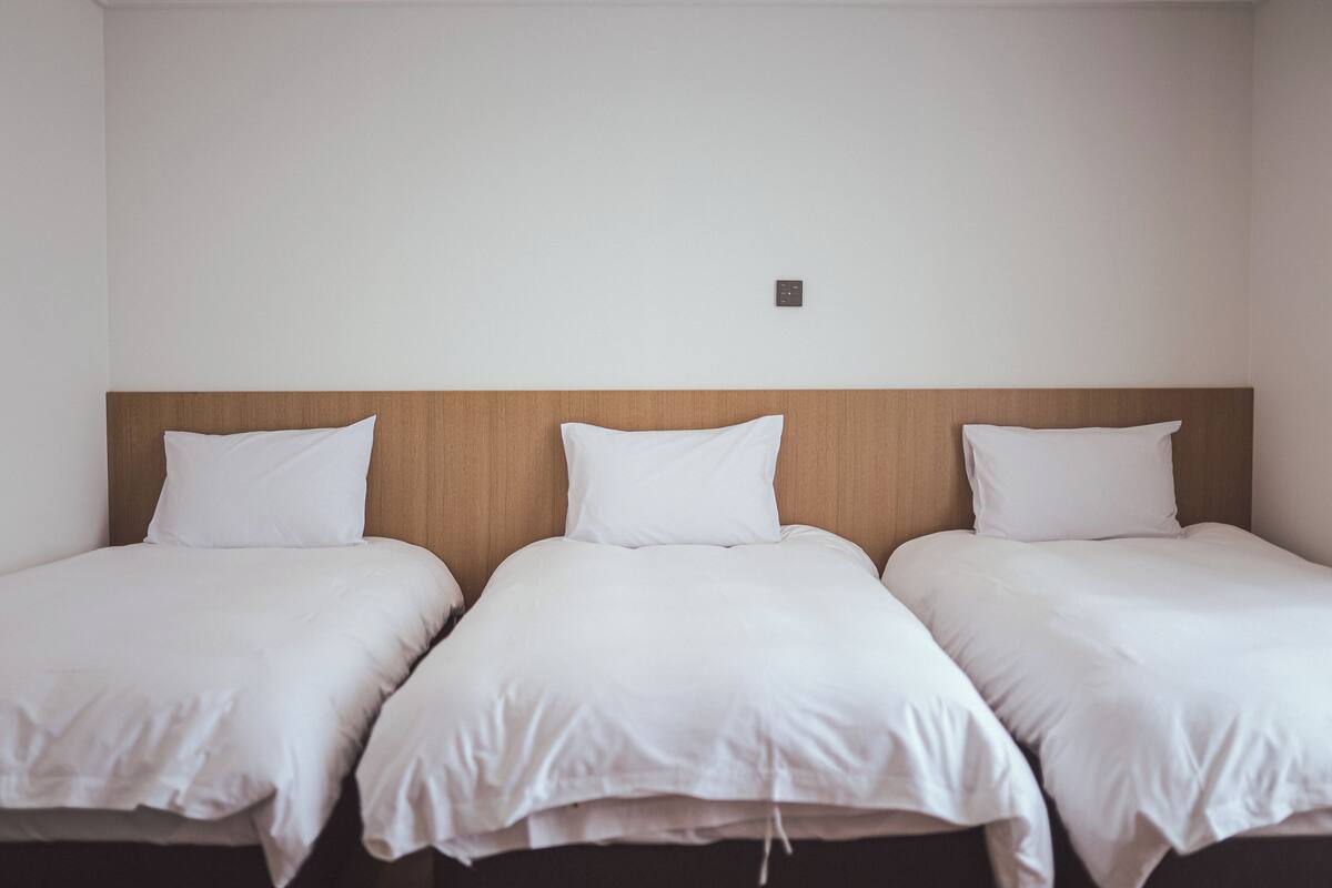 Ett hotellrum med tre separata sängar placerade längs en enkel vägg, alla med vita sängkläder och ett trägaveltavla.