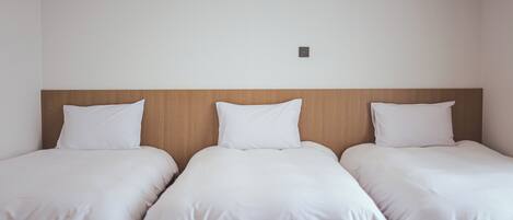 A hotel room with three single beds aligned against a plain wall, each with white bedding and a wooden headboard.