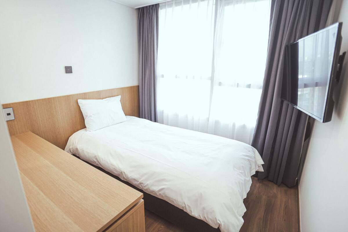 Ett modernt hotellrum med en säng, ett träbord vid sängen, en platt-TV som är monterad på väggen och ett fönster med gardiner.