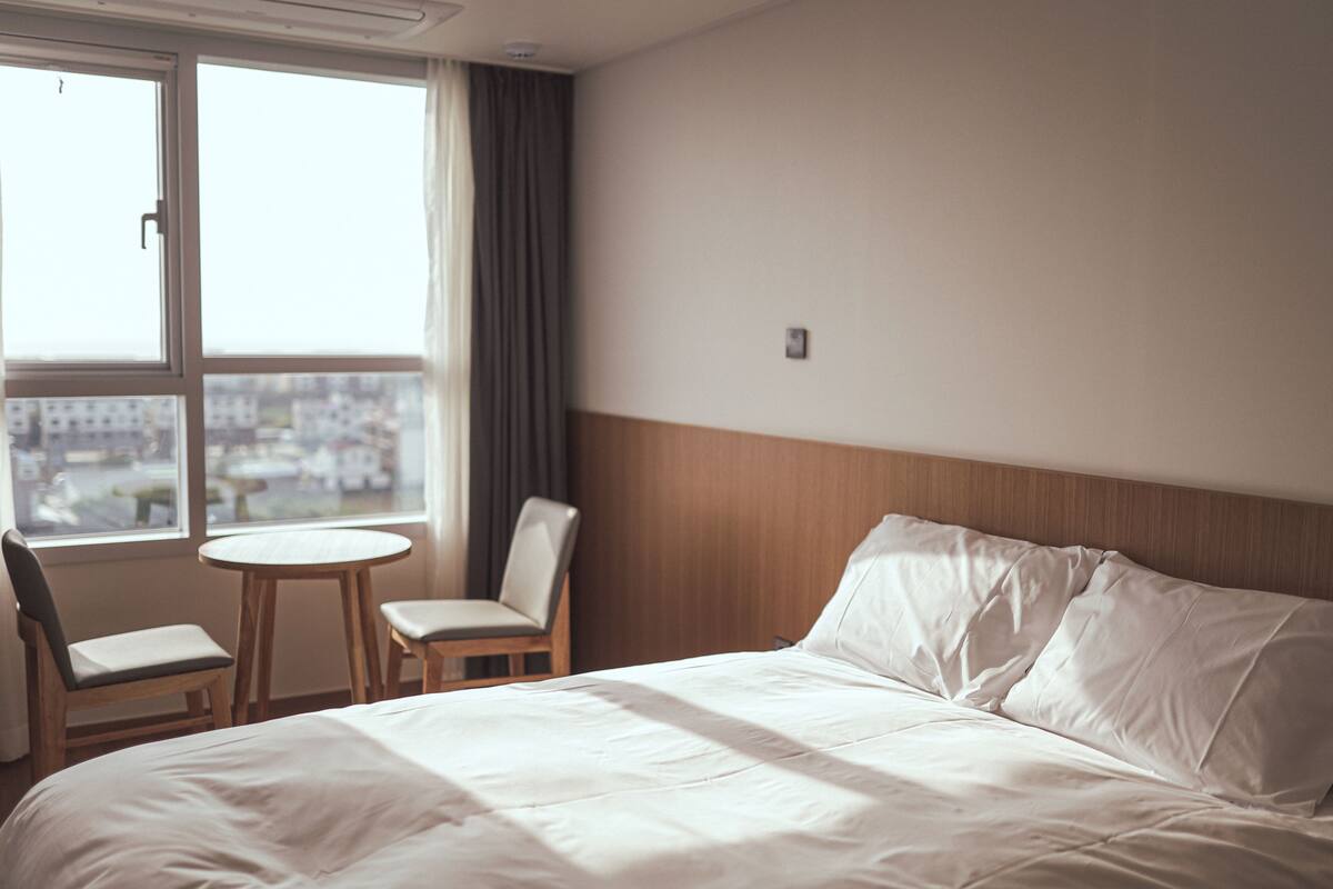 Ett hotellrum med en stor säng, ett litet runt bord, två stolar och utsikt över en stadssiluett genom ett stort fönster.