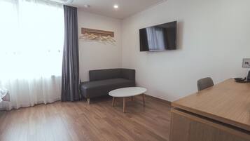 A modern room with a wooden floor, a grey sofa, a small round table, a TV mounted on the wall, and a wooden desk.