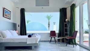 Habitación de hotel con cama, una silla, una mesita y vistas a palmeras y un edificio a través de amplios ventanales.