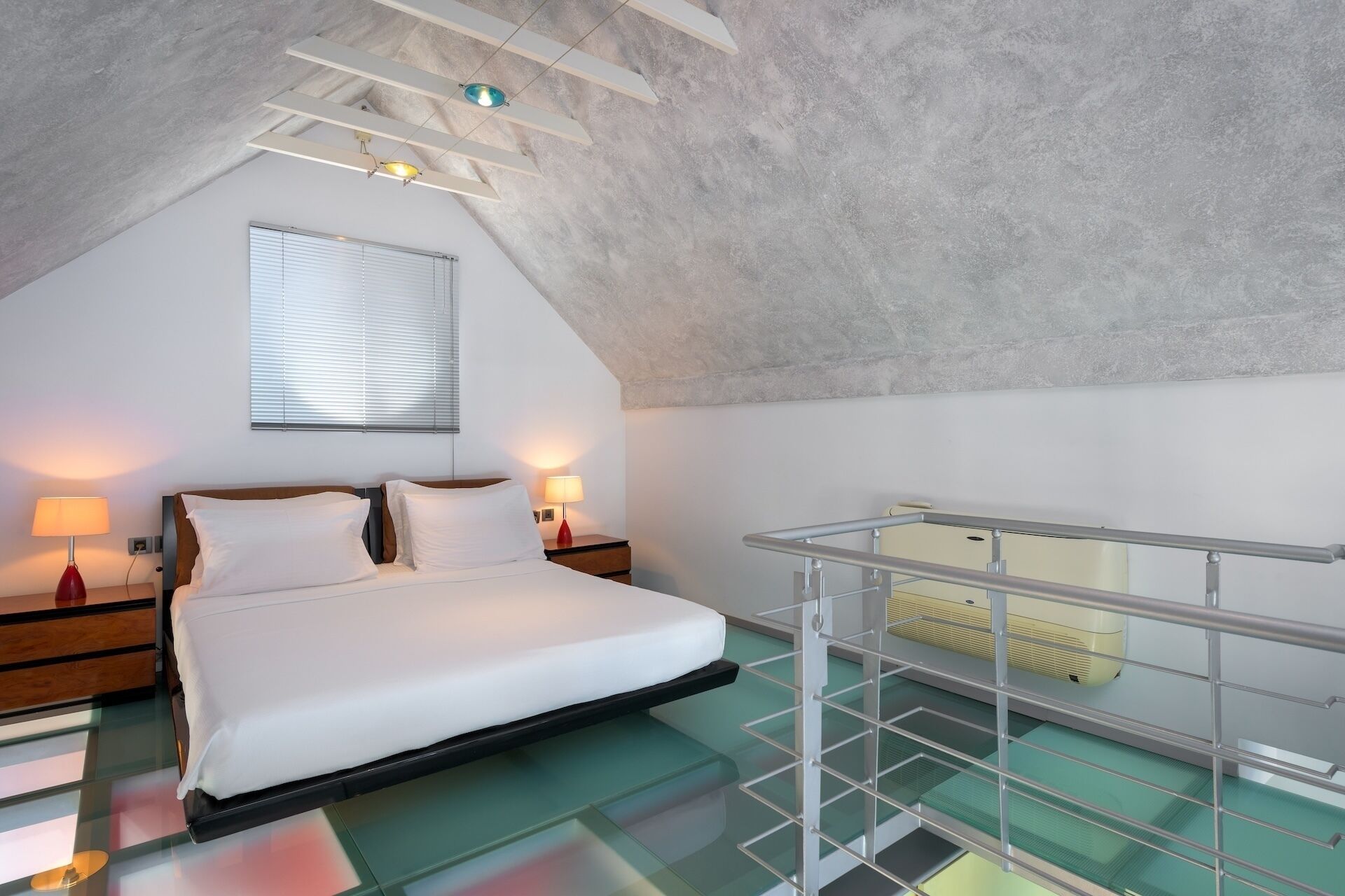 A modern bedroom with a glass floor, a bed with white linens, a wooden nightstand, and a ceiling with exposed beams and a skylight.