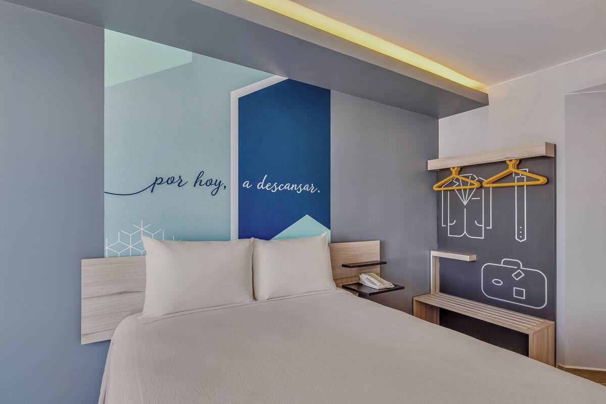 Une chambre d’hôtel moderne équipée d’un lit, de tables de chevet, d’un téléphone fixé au mur, et d’un mur orné d’un motif géométrique et d’une citation.
