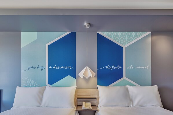 Moderni hotellihuone, jossa on sänky, valkoiset tyynyt, yöpöytä, jossa on puhelin, sekä geometriset seinätaideteokset, joissa on tekstiä.