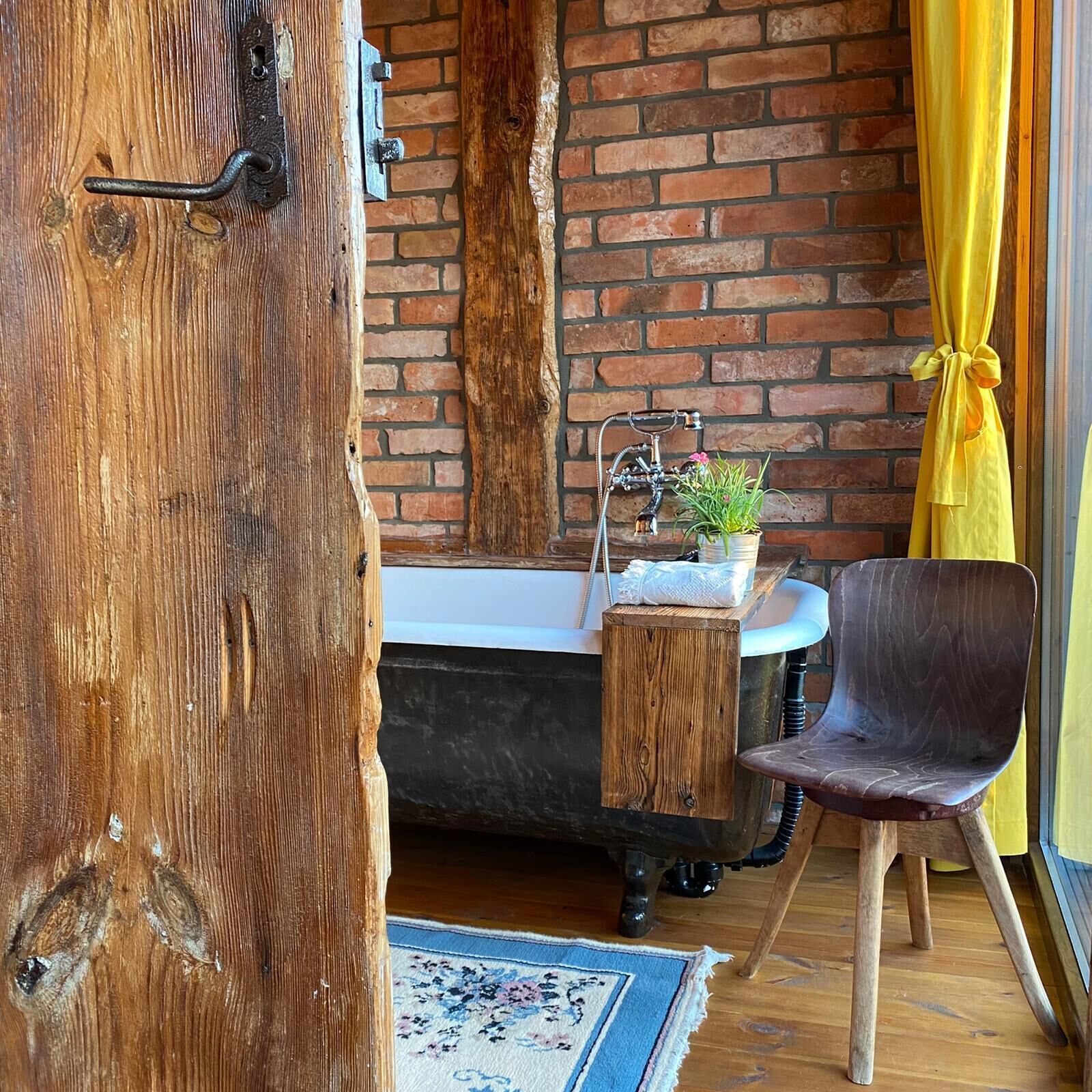 Ein rustikales Badezimmer mit einer Vintage-Badewanne auf Füßen, einem Holzschemel, einem Stuhl und einer Ziegelwand.