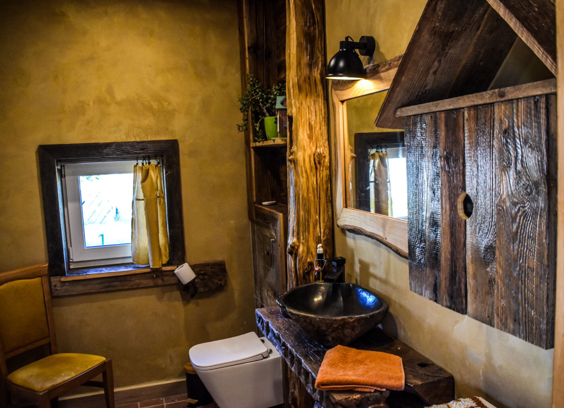 A rustic bathroom with a wooden vanity, a large mirror, a window with a curtain, a toilet, and a chair.
