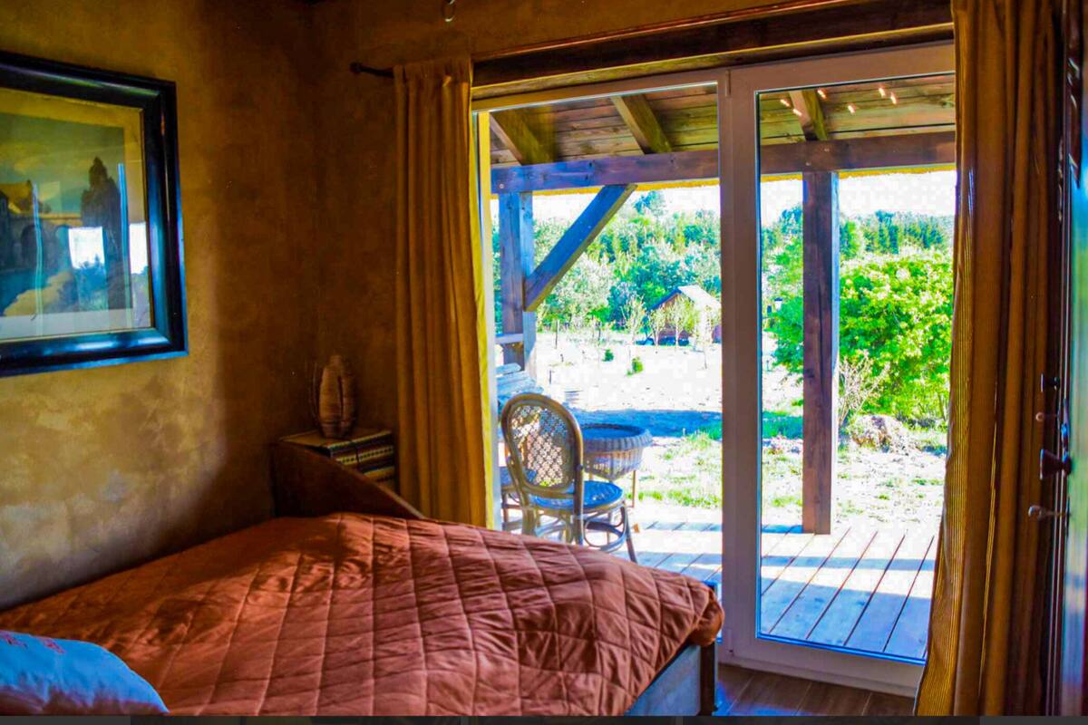 A bedroom with a bed, a chair, and a table near a sliding glass door leading to an outdoor area with a wooden deck and greenery.