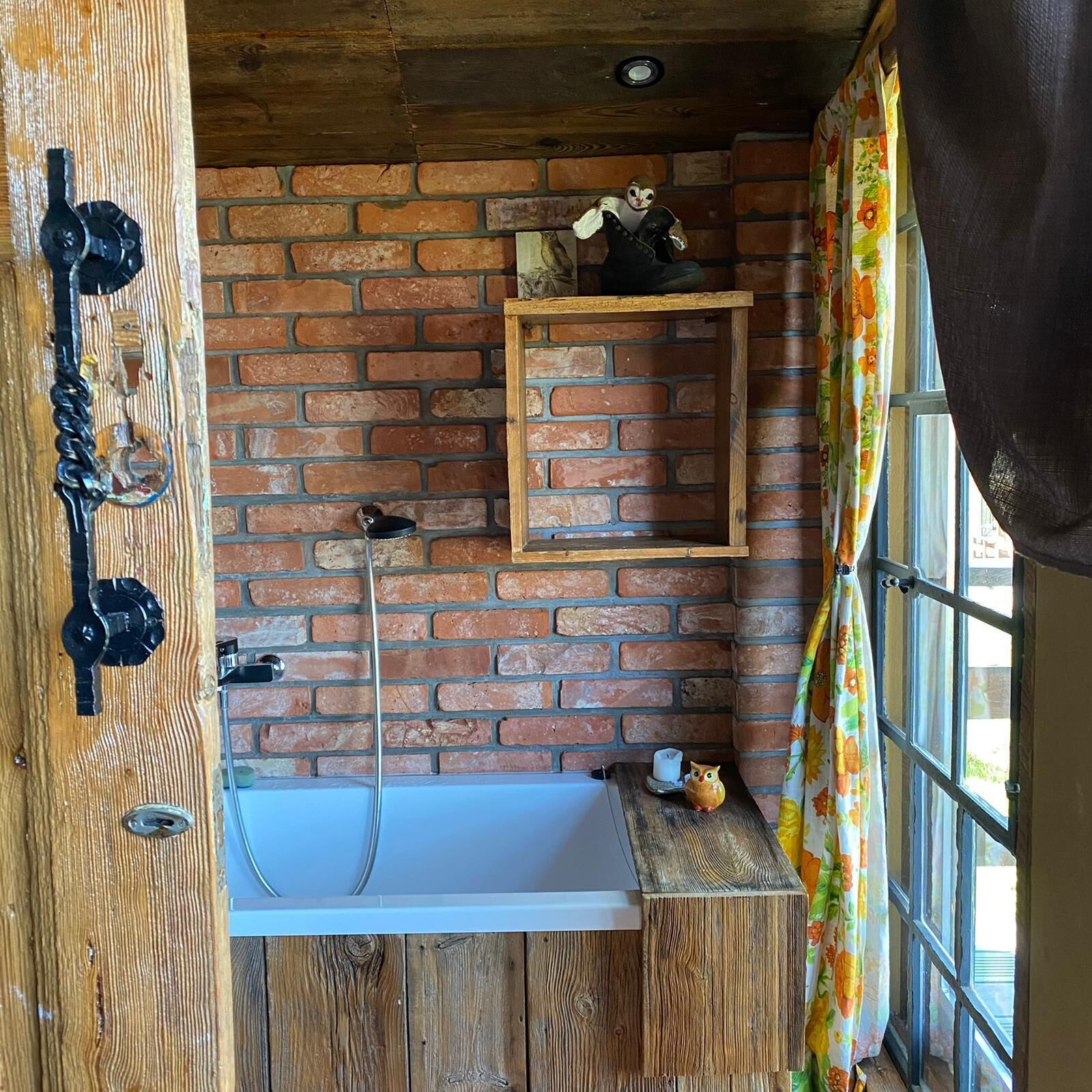 Une salle de bain rustique avec un mur en briques, une étagère en bois, une baignoire et une fenêtre ornée de rideaux à motifs floraux.