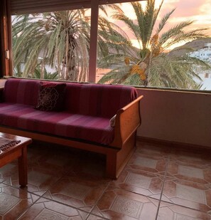 A room with a sofa and a wooden table, a tiled floor, and a view of palm trees and a building through a window.